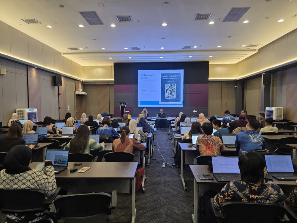 Training room with rows of attendees on laptops facing two projector screens that display a pre-assessment question slide and a QR code.